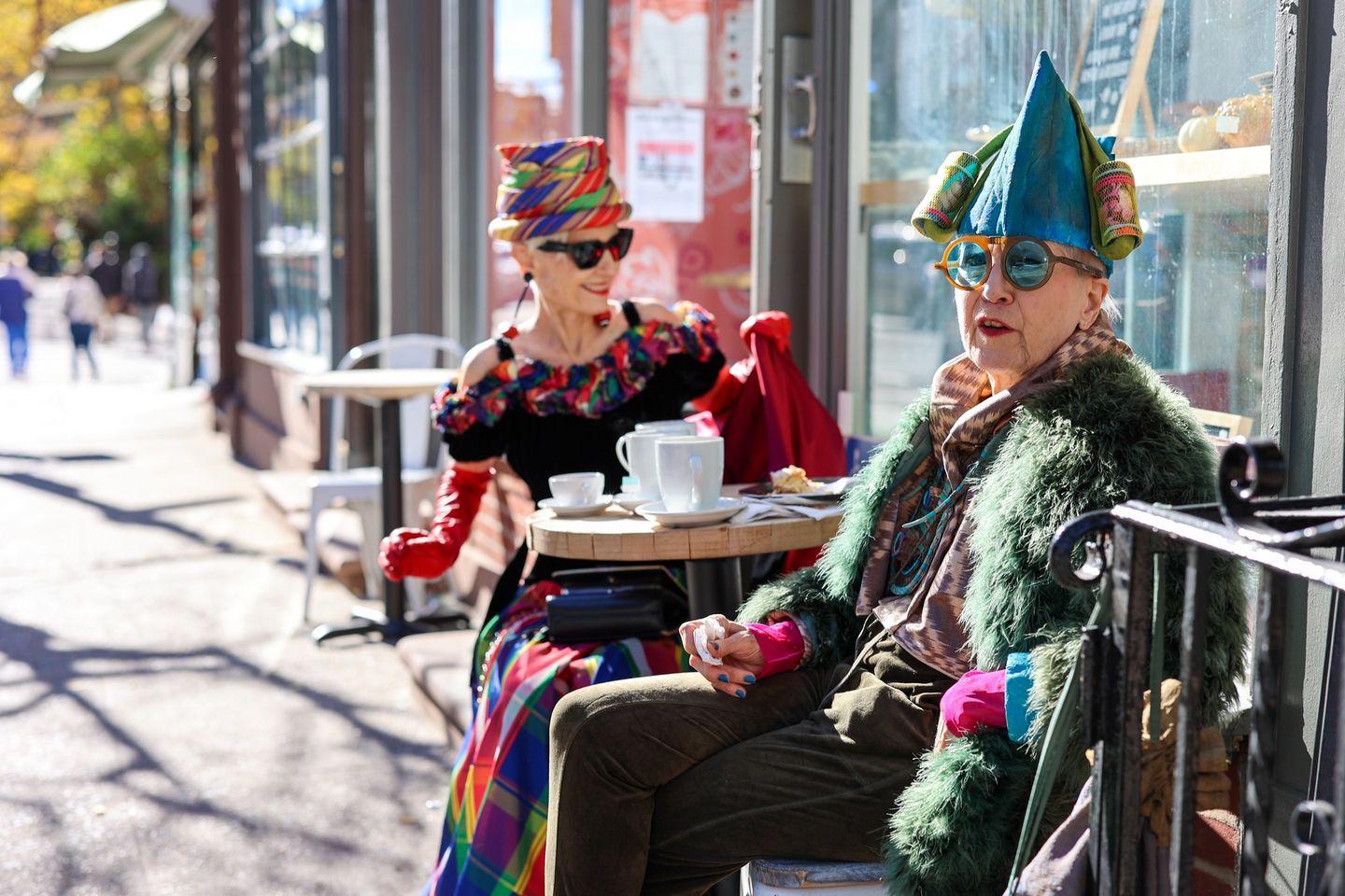 Two women at outdoor café dressed in colorful garb and hats