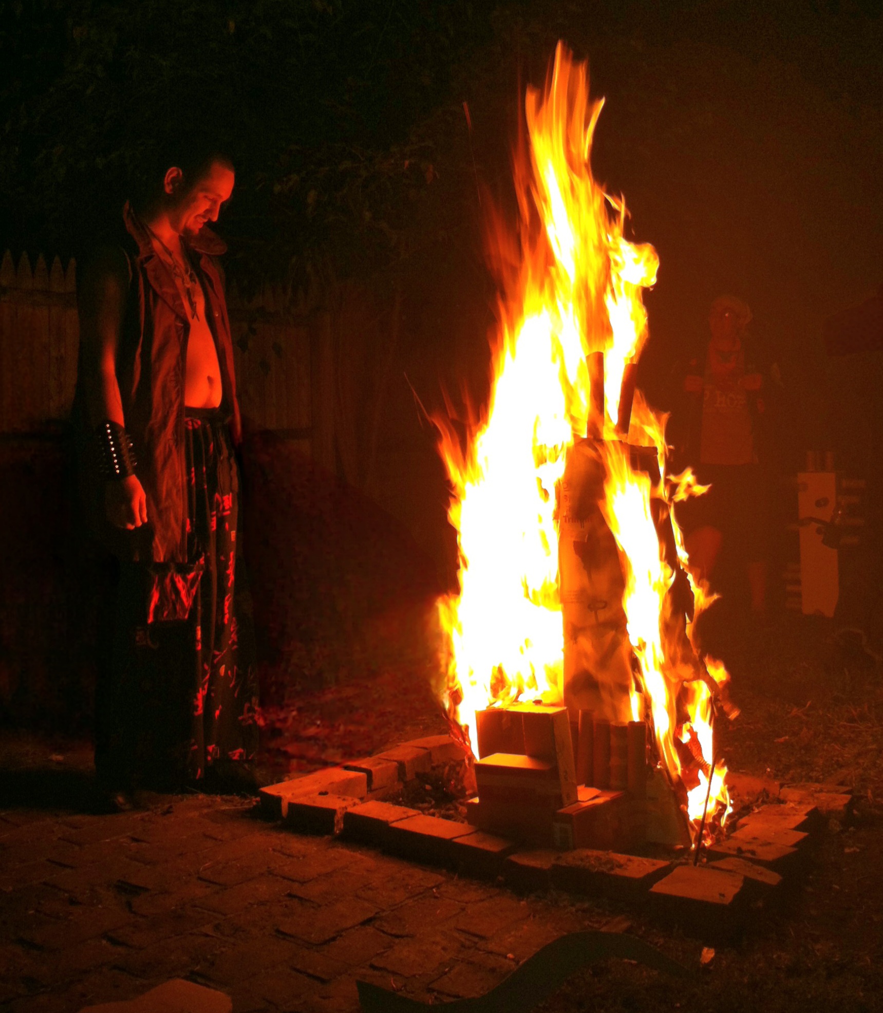 man in open shirt and flowing pants staring into the fire of a burning box