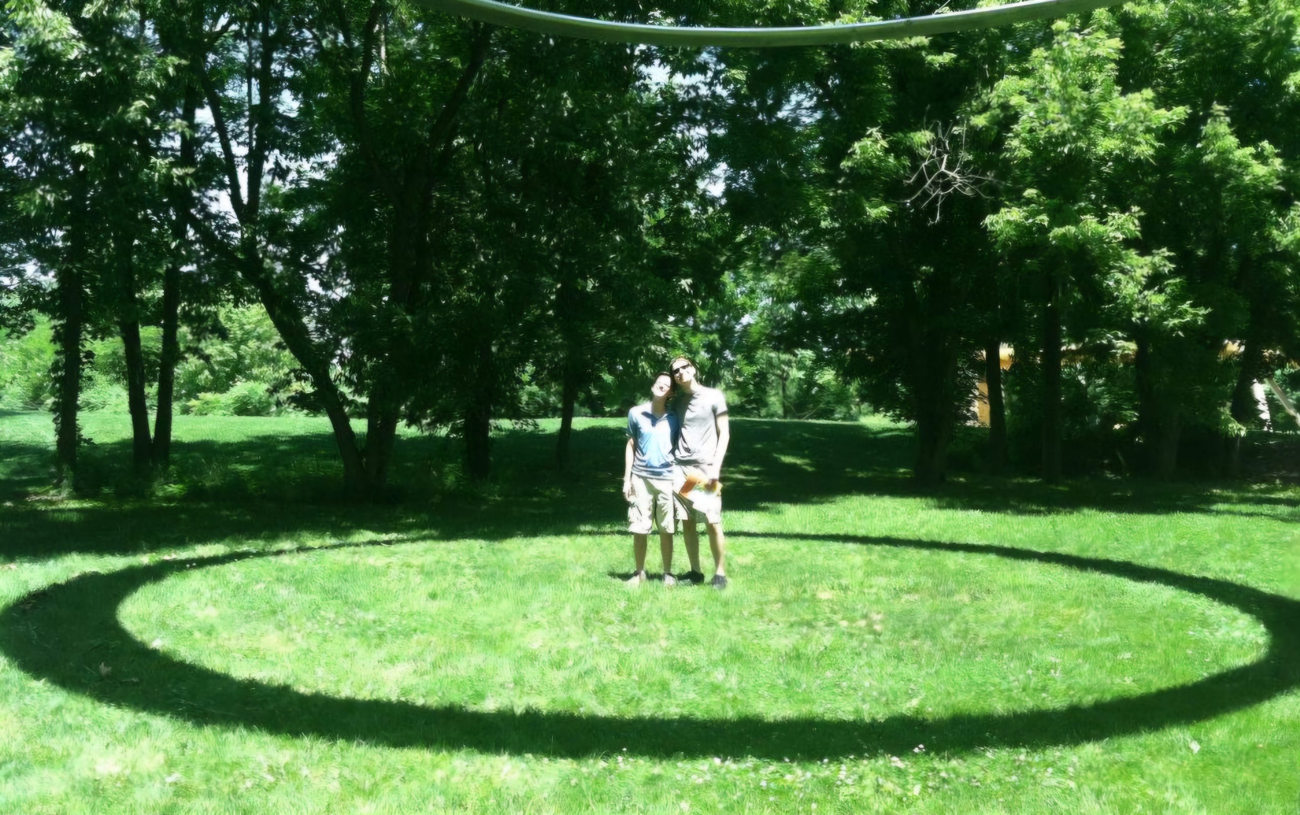 Two men with arms around one another standing in the grass with a circular shadow around them on the ground