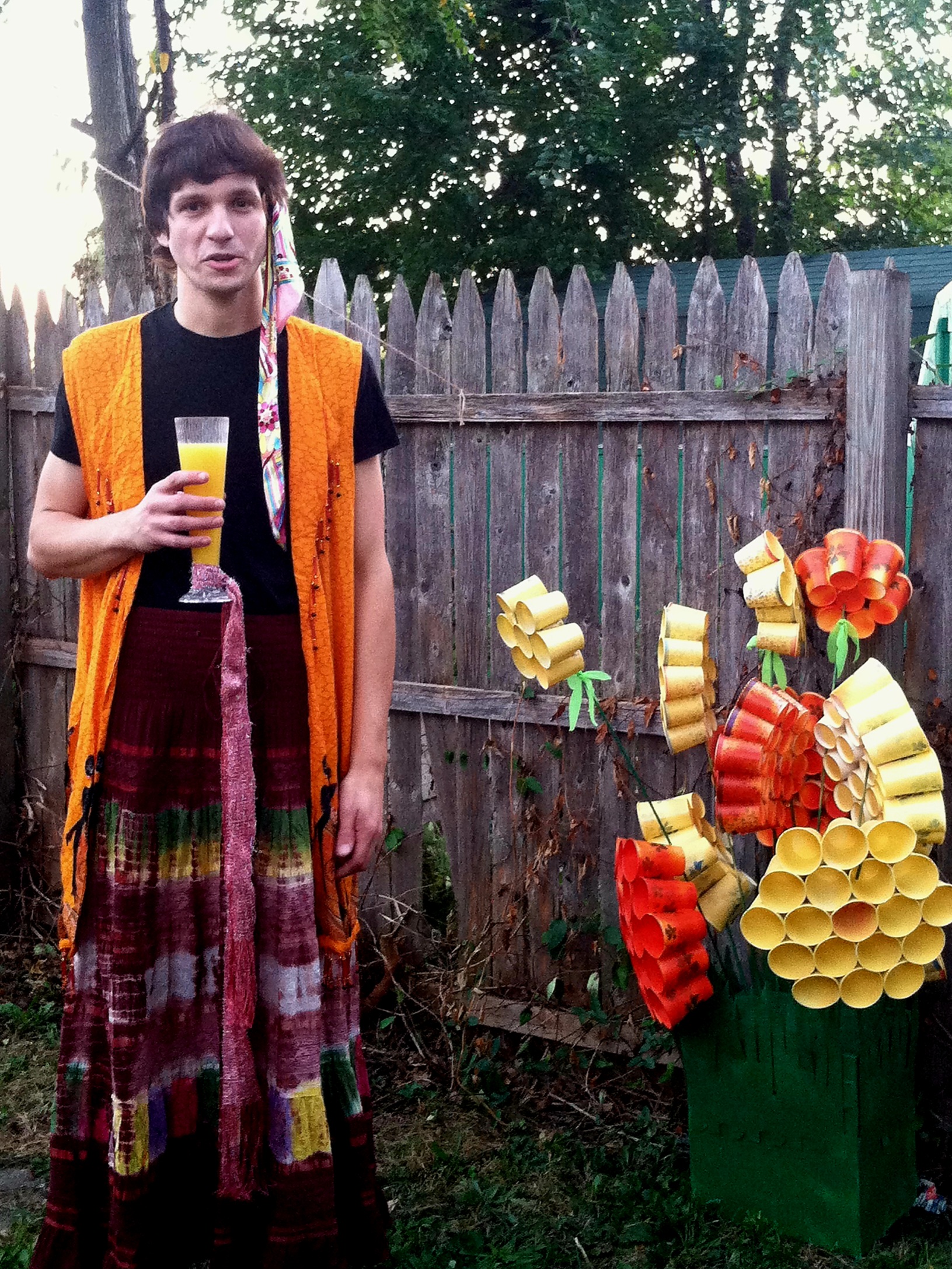 Man in a colorful outfit with head scarg and skirt holding a drink next to some flowers made out of paper cups