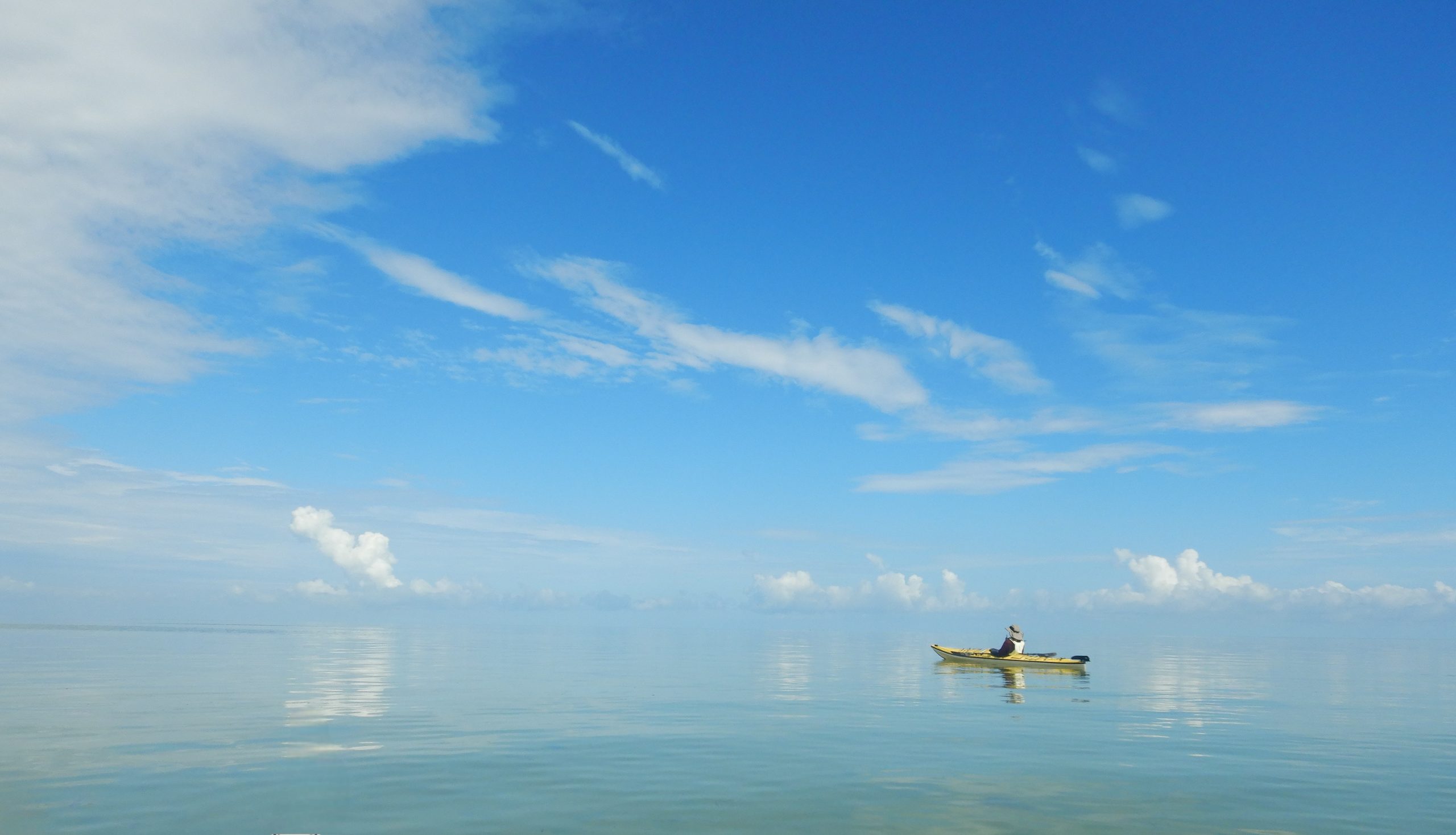 Photo of a sea kayak in the distance on an ocean with a blue sky and white clouds. The water is so calm, it's difficult to tell where the water ends and sky begins.