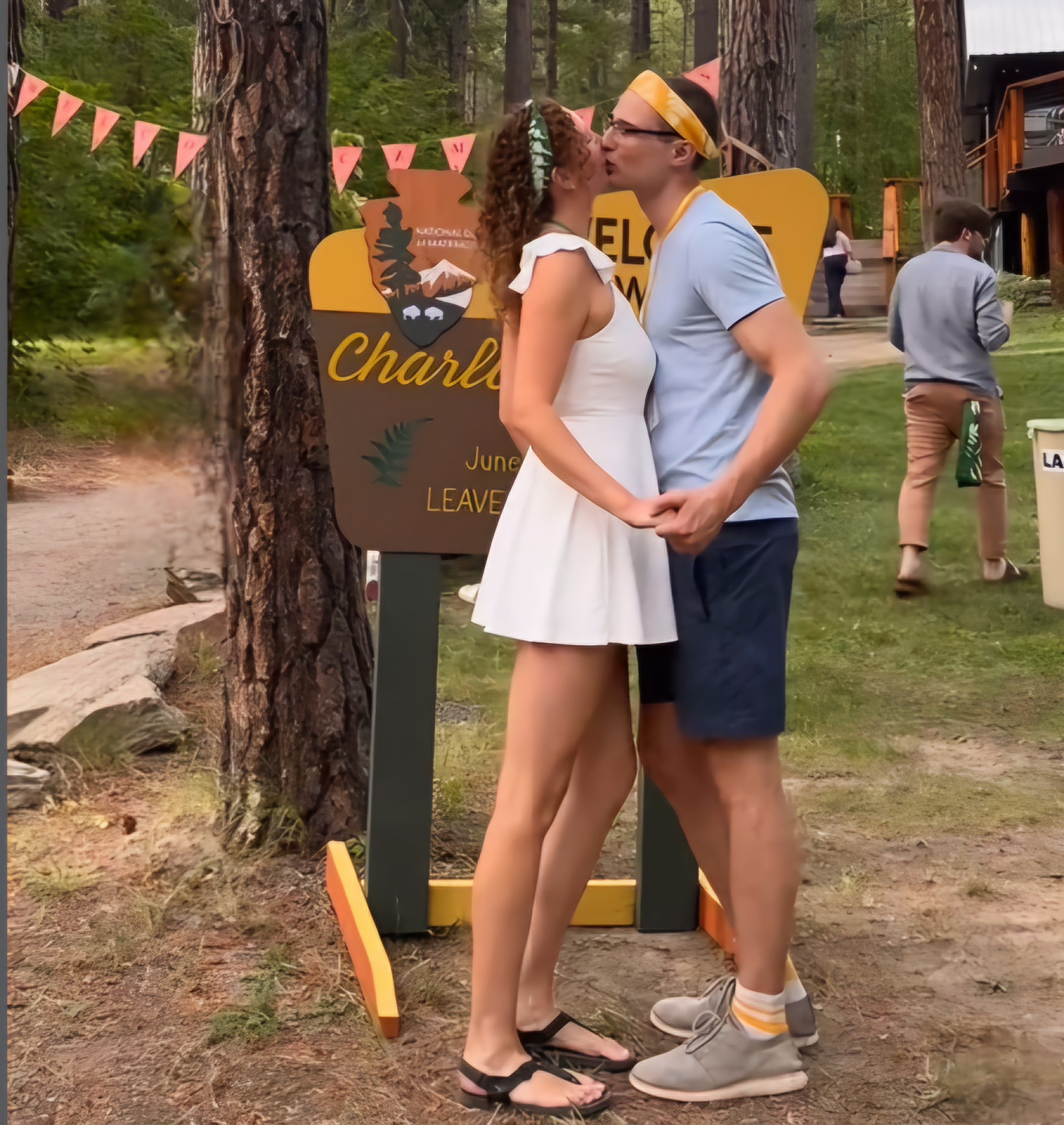 Couple kissing in an park. She has on a short, simple wedding dress. He is dressed in t-shirt and shorts with a bandana tied around his head.