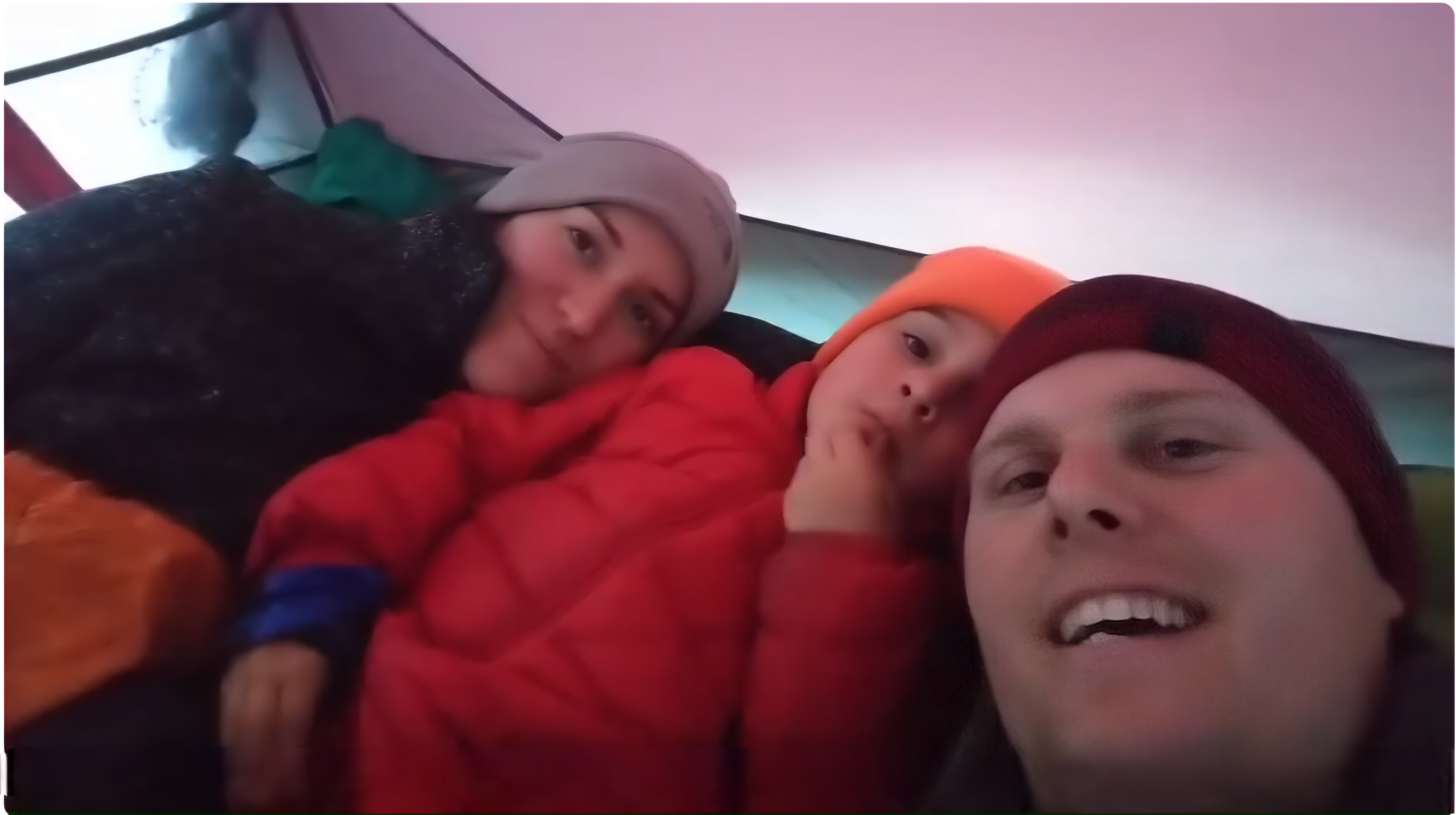 Young mom, son, and dad in a tent wearing puffy coats and knit hats laying on sleeping bags taking a selfie.