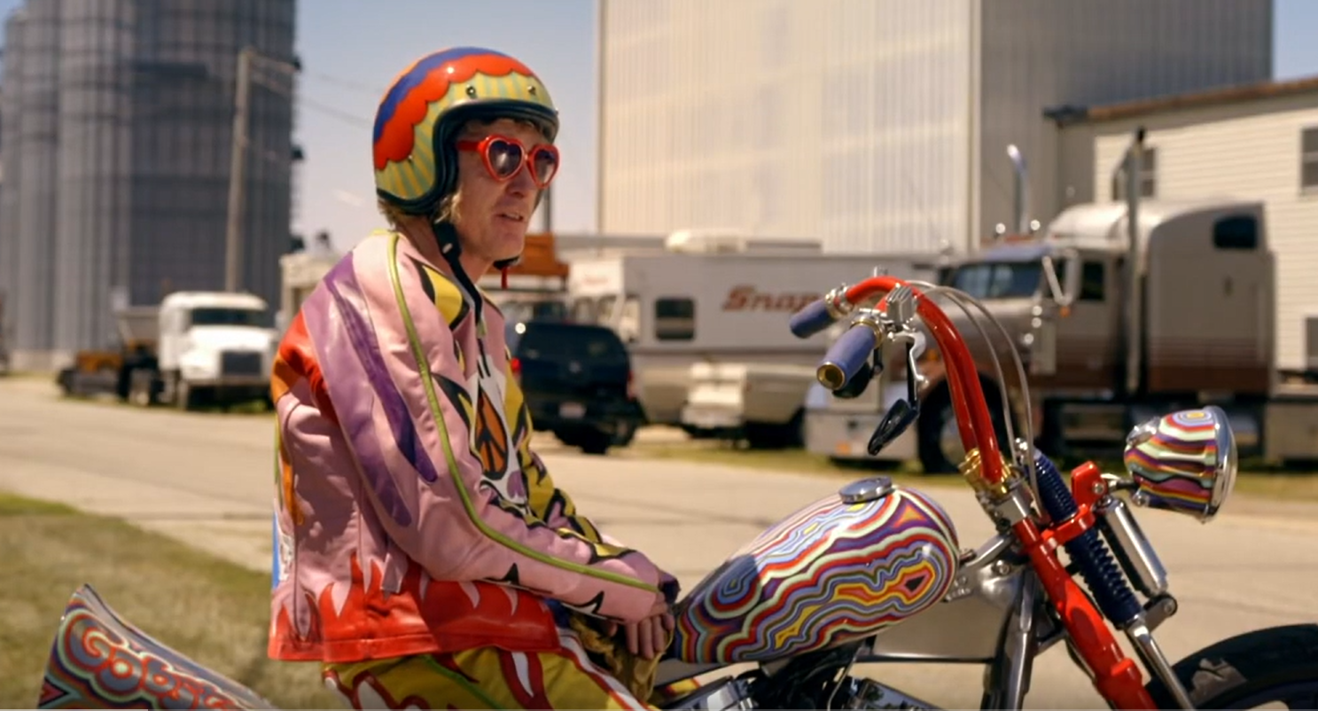 Grayson Perry on a motorcycle with bright colors and swirls wearing an equally decorative and bright leather jacket, pants, helmet, and heart-shaped sun glasses