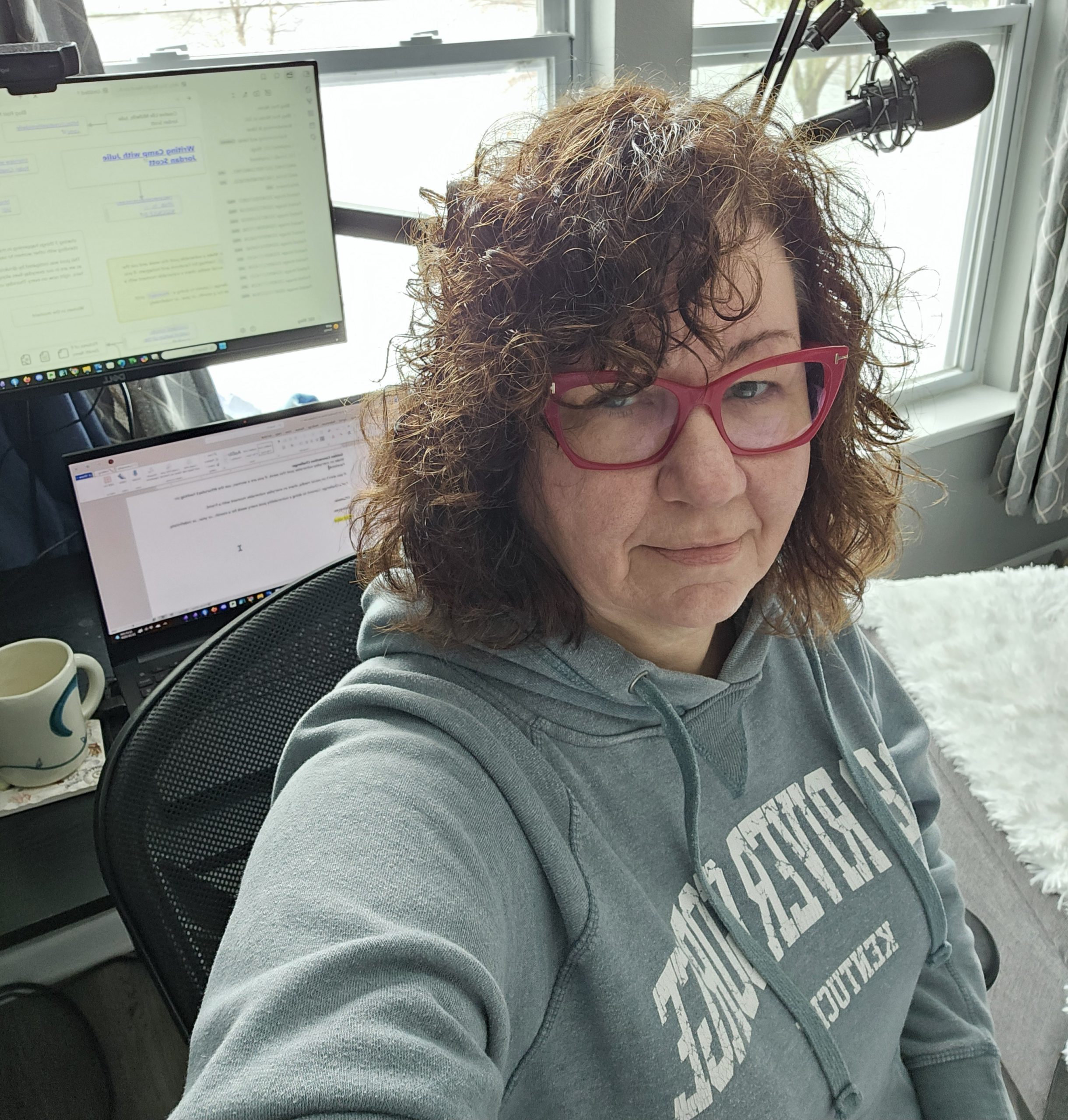 Selfie of the author in a Red River Gorge sweatshirt at her desk with window and computer monitors in the background