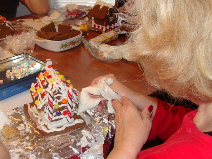 Older woman from the back making a gingerbread house at a kitchen table full of supplies