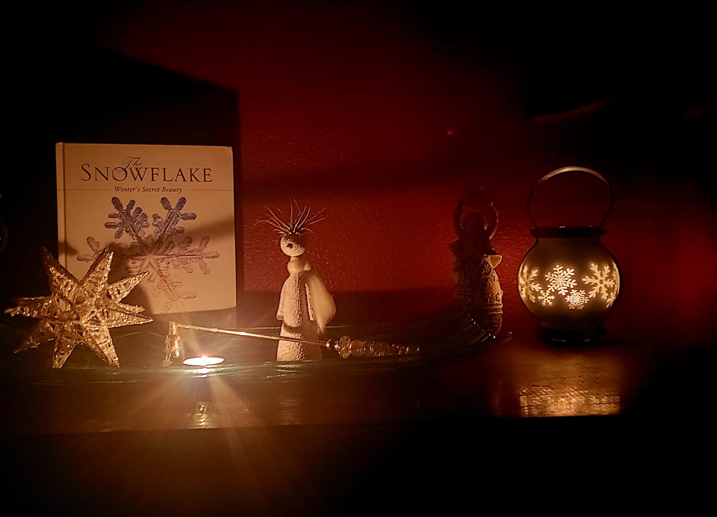 Candle-lit scene of Christmas decorations, a snowflake book, Moravian star,  snowflake lit globe, and angel.