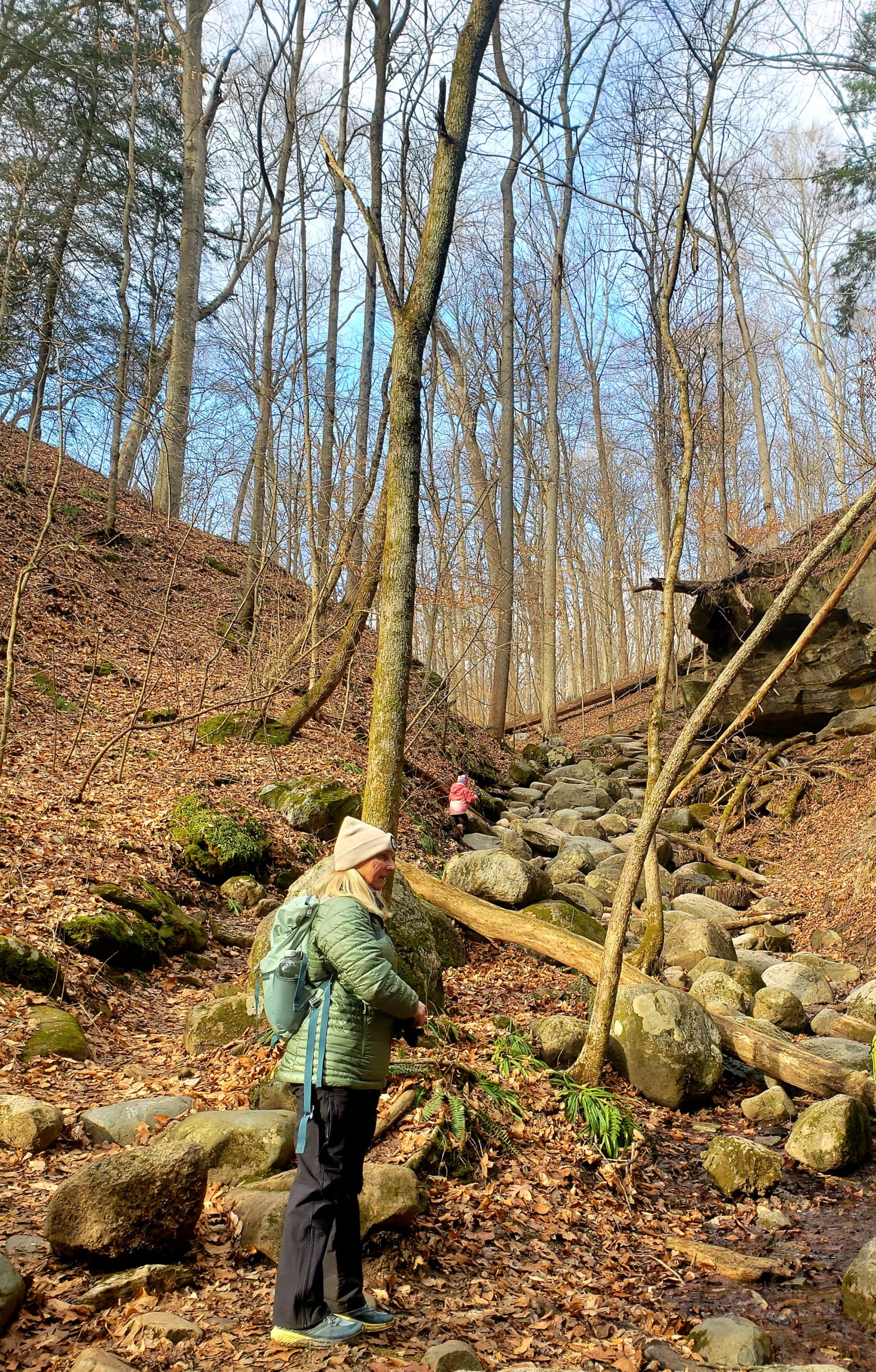 Child in background climbing up a boulder scramble on a trail in winter. Adult standing in foreground.